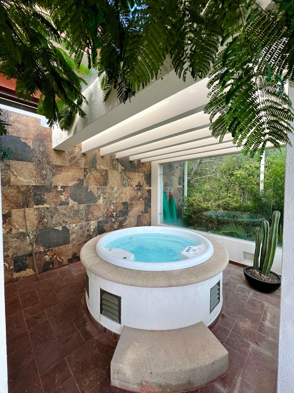 A private jacuzzi is situated within a serene space, surrounded by natural stone walls and greenery. Large windows provide a view of the outdoor plants, while a patterned floor adds texture. Ferns hang from the ceiling, enhancing the tranquil ambiance.