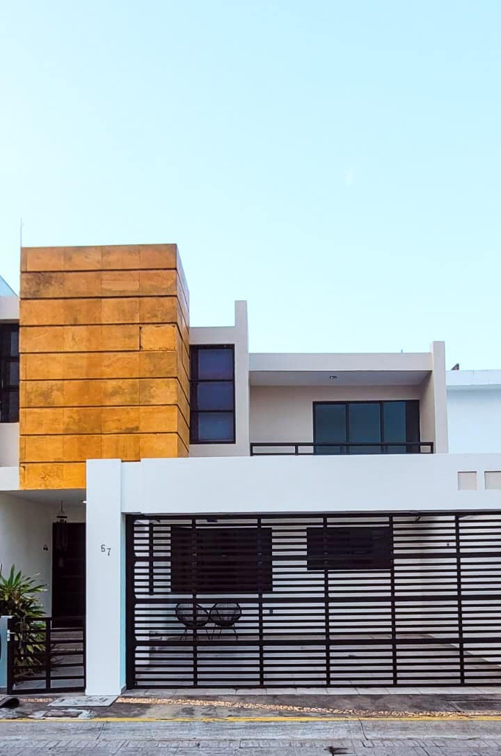 A modern house exterior features a striking gold accent wall, complemented by large windows and a black metal gate. A small seating area is visible in front, surrounded by minimal landscaping. The home’s design reflects a contemporary aesthetic, blending style with functionality.
