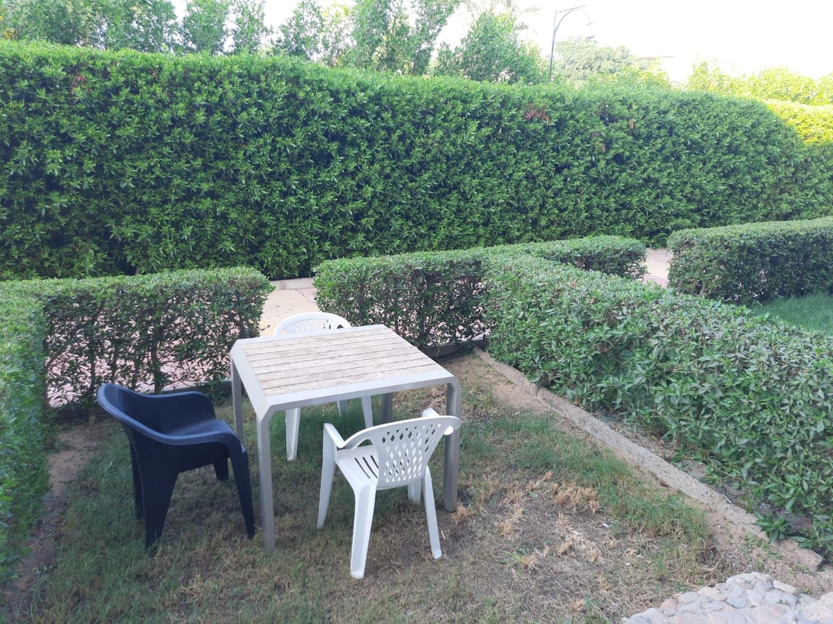 An outdoor seating area features a wooden table surrounded by three chairs, including one black and two white plastic options. Lush green hedges provide privacy, framing the space with neatly trimmed foliage.