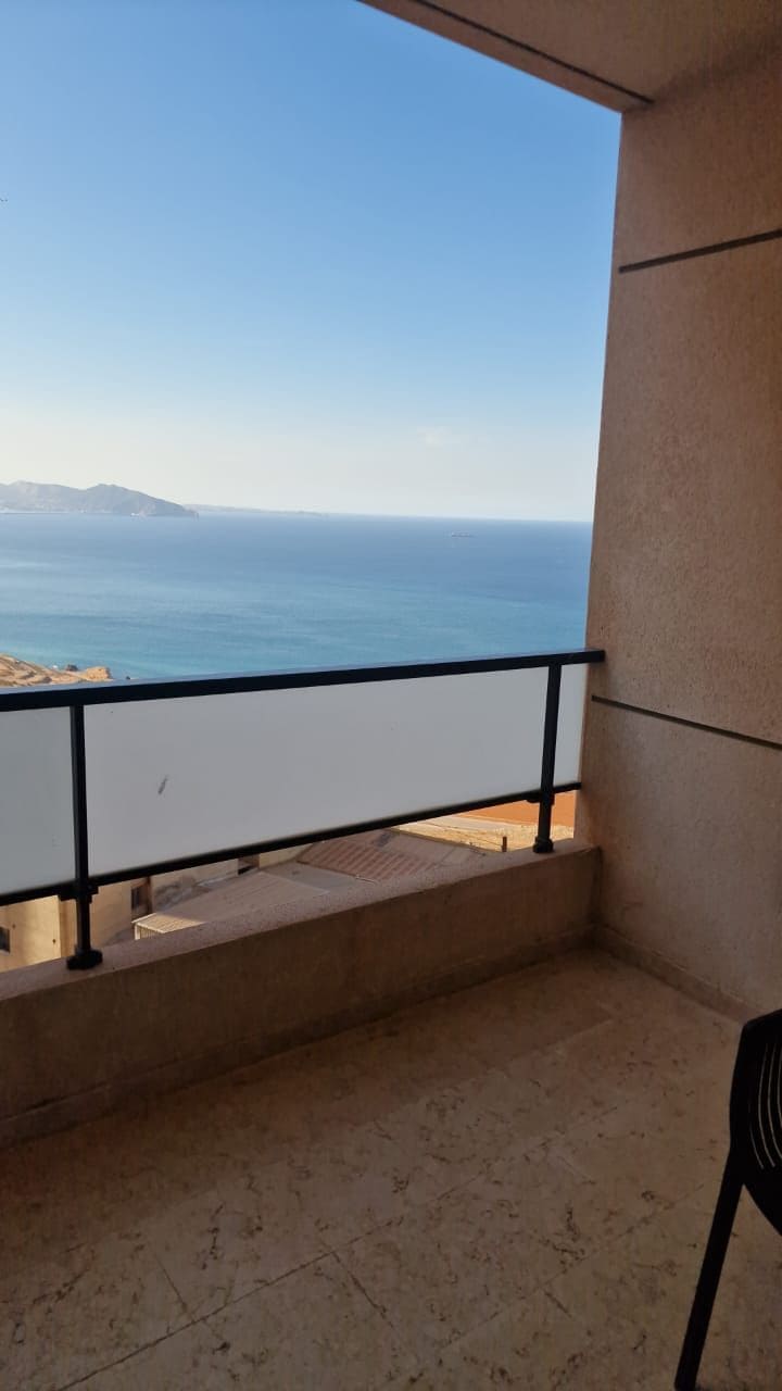 A spacious balcony offers a stunning view of the sea, framed by a minimalistic railing. The calm blue water extends toward distant mountains under a clear sky. The area is finished with a smooth stone floor, suggesting a tranquil outdoor space for relaxation.
