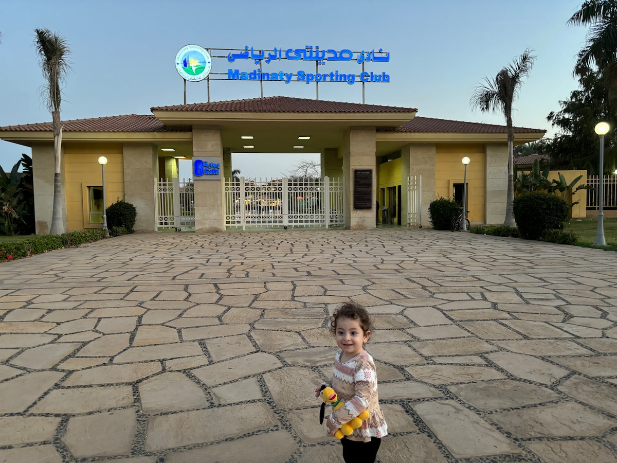 The entrance of Madinaty Sporting Club is showcased, featuring a well-maintained facade with landscaped areas. A smooth stone pathway leads to a gated entry. Soft exterior lighting enhances the inviting atmosphere as palm trees frame the structure against the dusk sky.