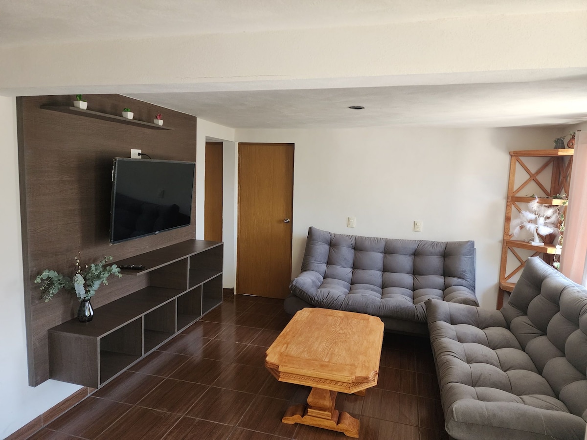 A welcoming living area features a cozy sectional sofa arranged around a natural wood coffee table. A television is mounted on the wall above a sleek brown media console, complemented by decorative accents. Natural light fills the space, enhancing its uncluttered design.