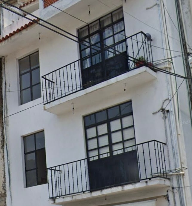 A three-story building is shown, featuring a clean white façade. Each floor includes large windows with black frames, enhancing natural light penetration. Two balconies with decorative railings are present, adorned with potted plants, offering an outdoor space for relaxation.
