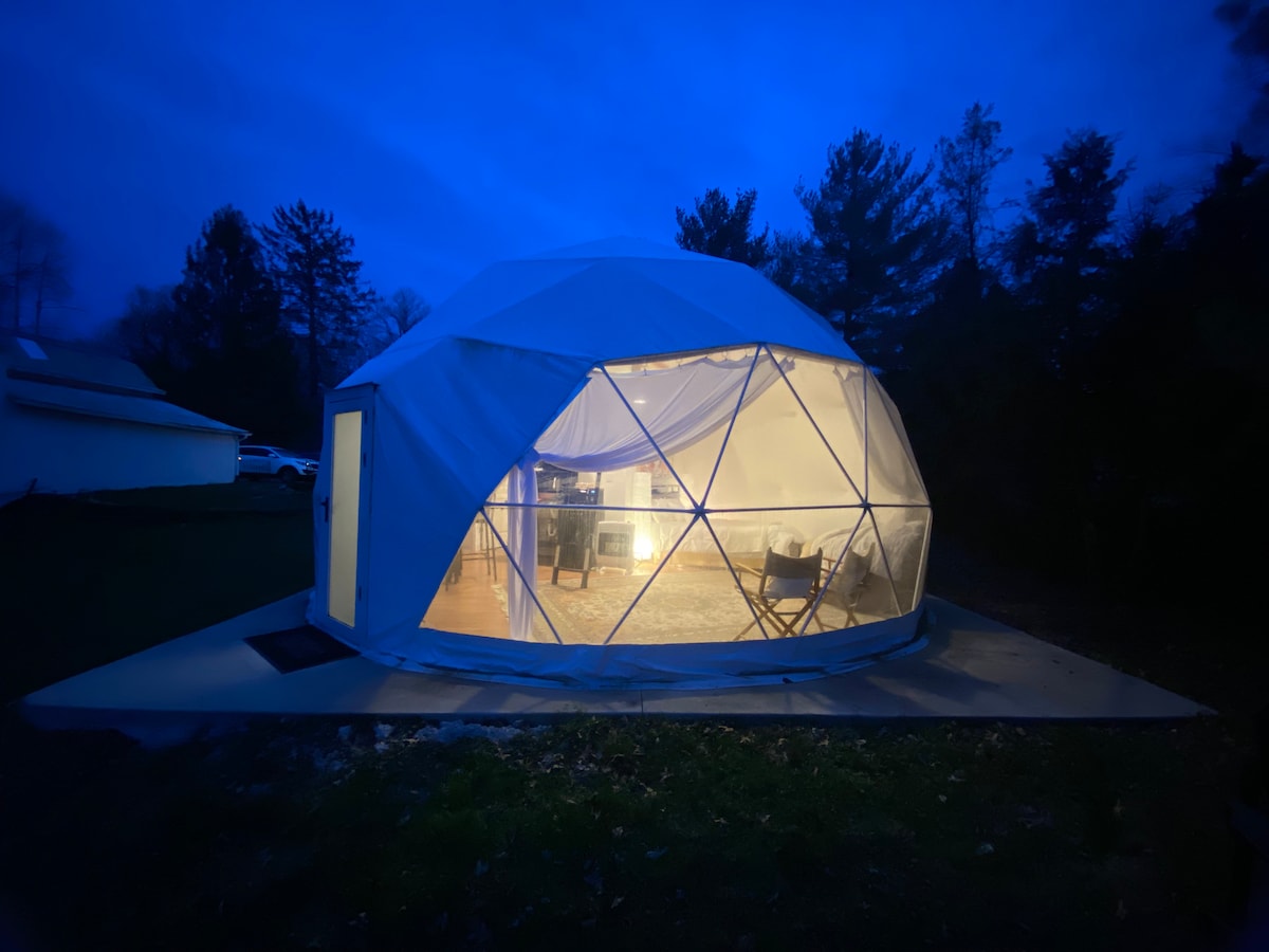 An illuminated geodesic dome is shown at night, featuring transparent walls that reveal a cozy interior space. A patio area is visible, leading to a door that opens into the dome. Surrounding trees frame the serene setting, enhancing the peaceful atmosphere.