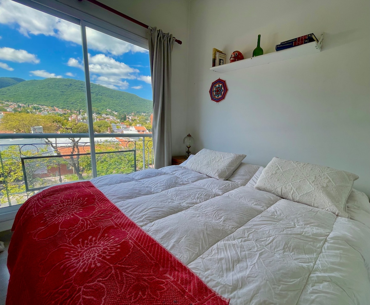A spacious sleeping area features a large, comfortable bed adorned with white bedding and a vibrant red throw. Natural light floods in through the floor-to-ceiling window, which offers a scenic view of the surrounding mountains and cityscape.