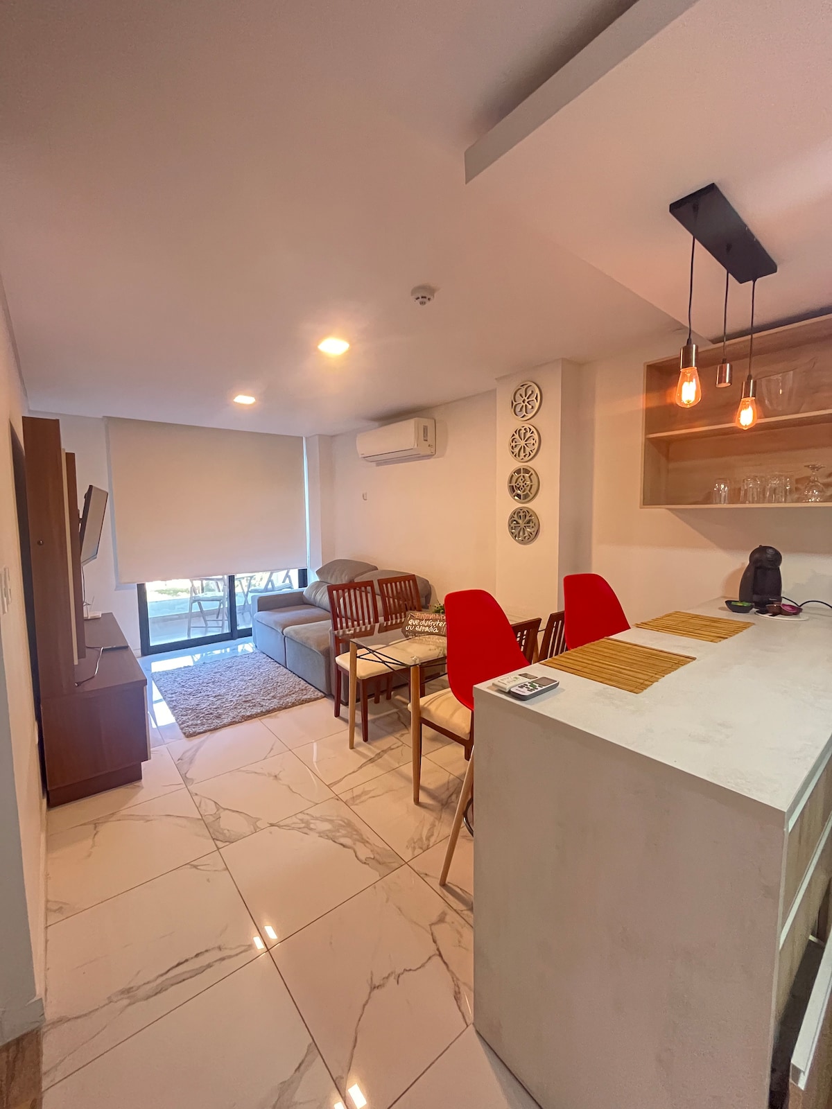 The living area features a sofa, accompanied by two wooden chairs and a dining table set for four. A television is placed against the wall, while light enters through the large window, enhancing the bright and open feel of the space.