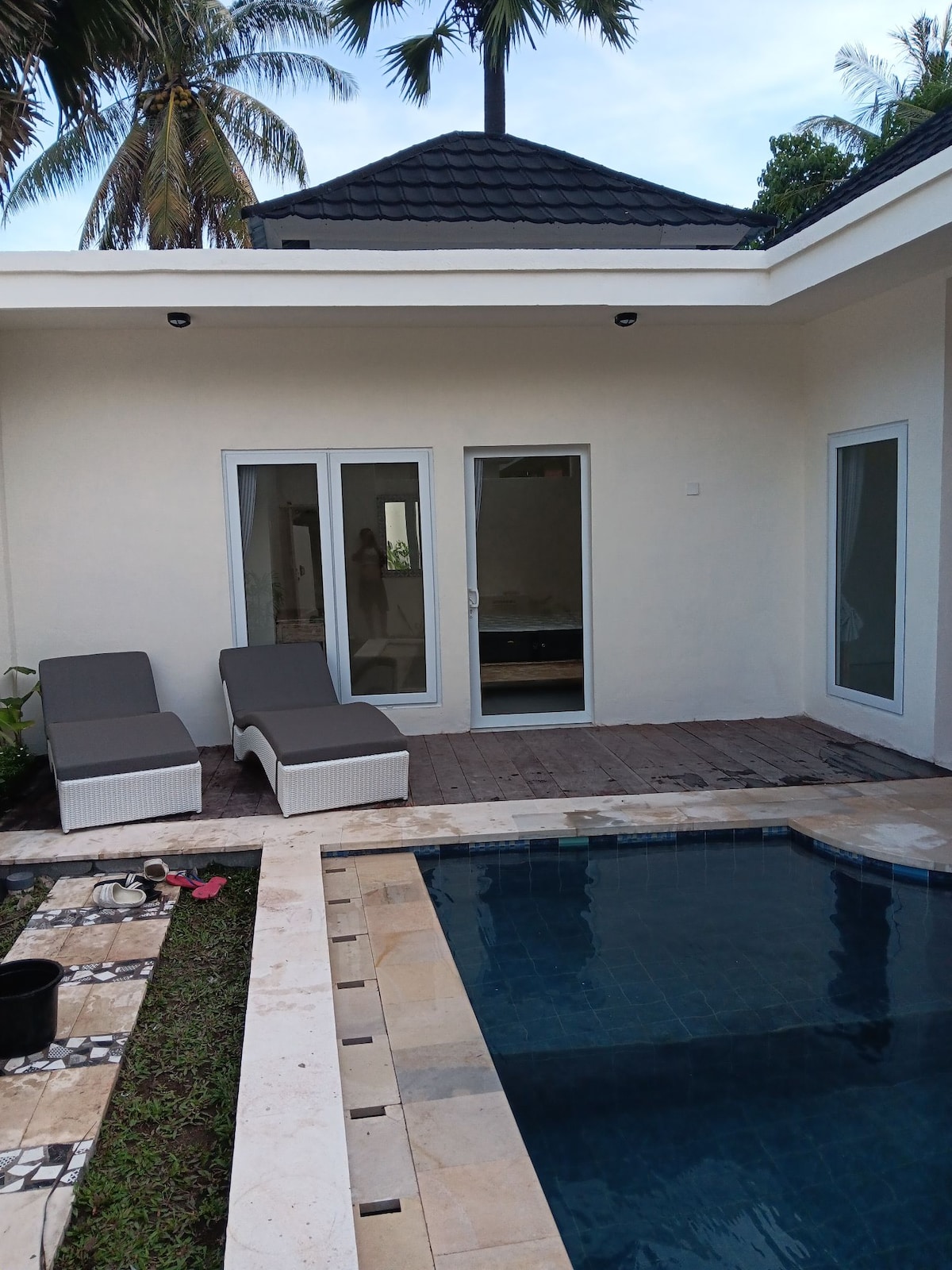 An outdoor area features a private pool surrounded by a tiled deck. Two loungers are positioned beside the pool, with a backdrop of palm trees and a simple structure. Large glass doors provide access to the interior space, adding a sense of openness.