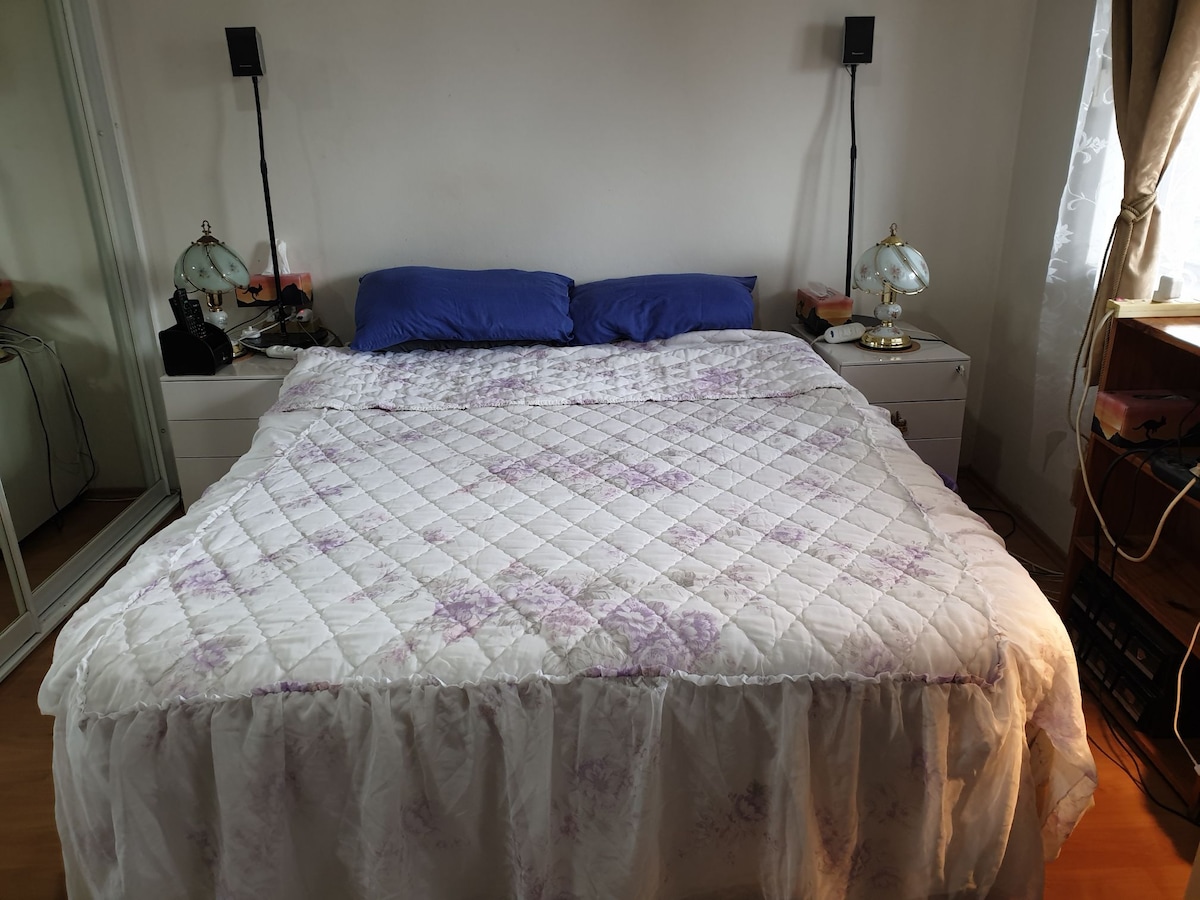 A spacious bed is centered in the room, featuring a white quilted cover with subtle purple floral patterns. Two bedside lamps are positioned on either side, providing a soft glow. A mirrored closet and a window with light curtains are visible in the background.