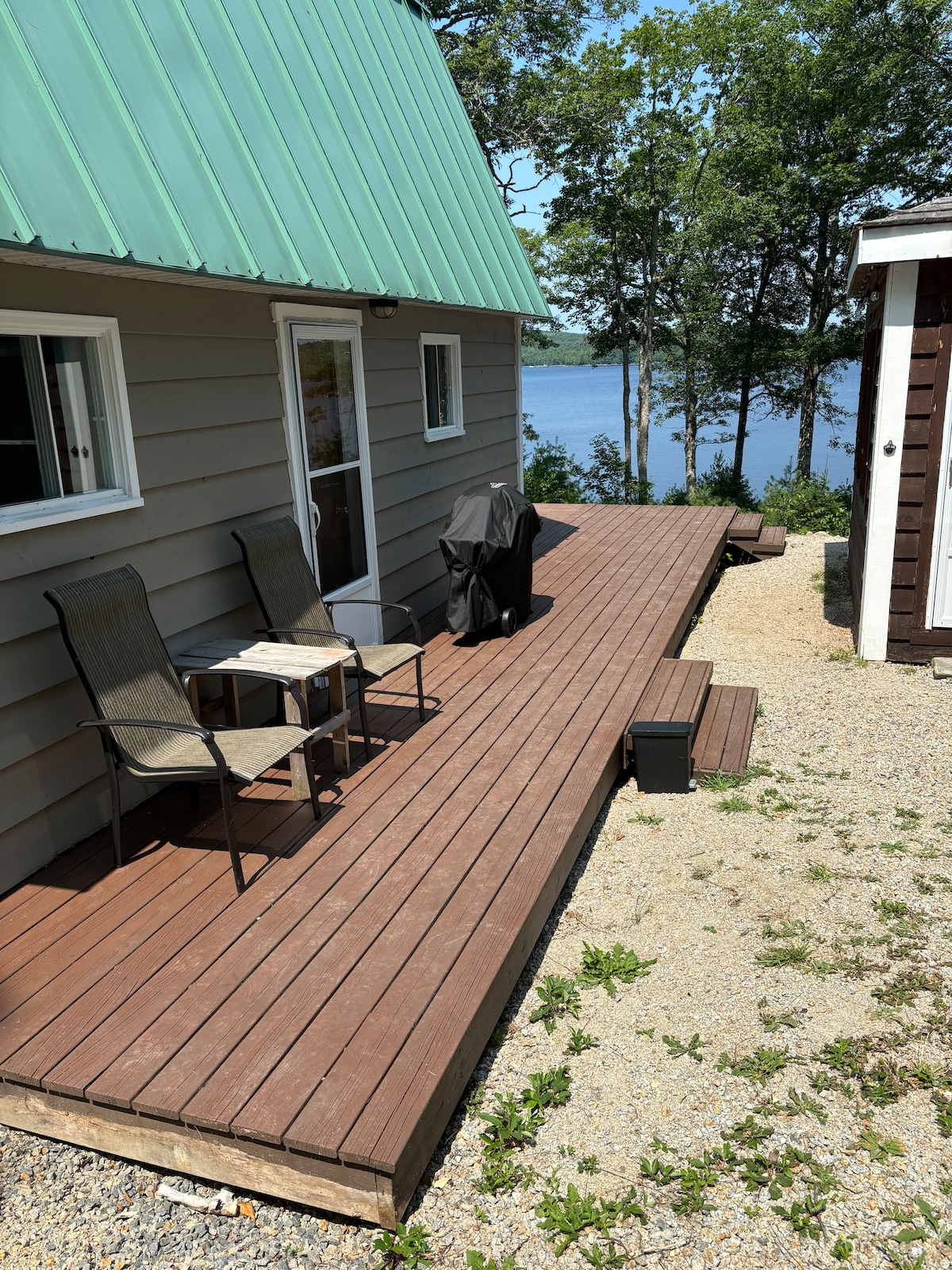 A wooden deck extends from the cottage, featuring two chairs and a small table. A barbecue grill is positioned on the deck, and the view of the lake is visible in the background. Surrounding greenery enhances the natural setting.
