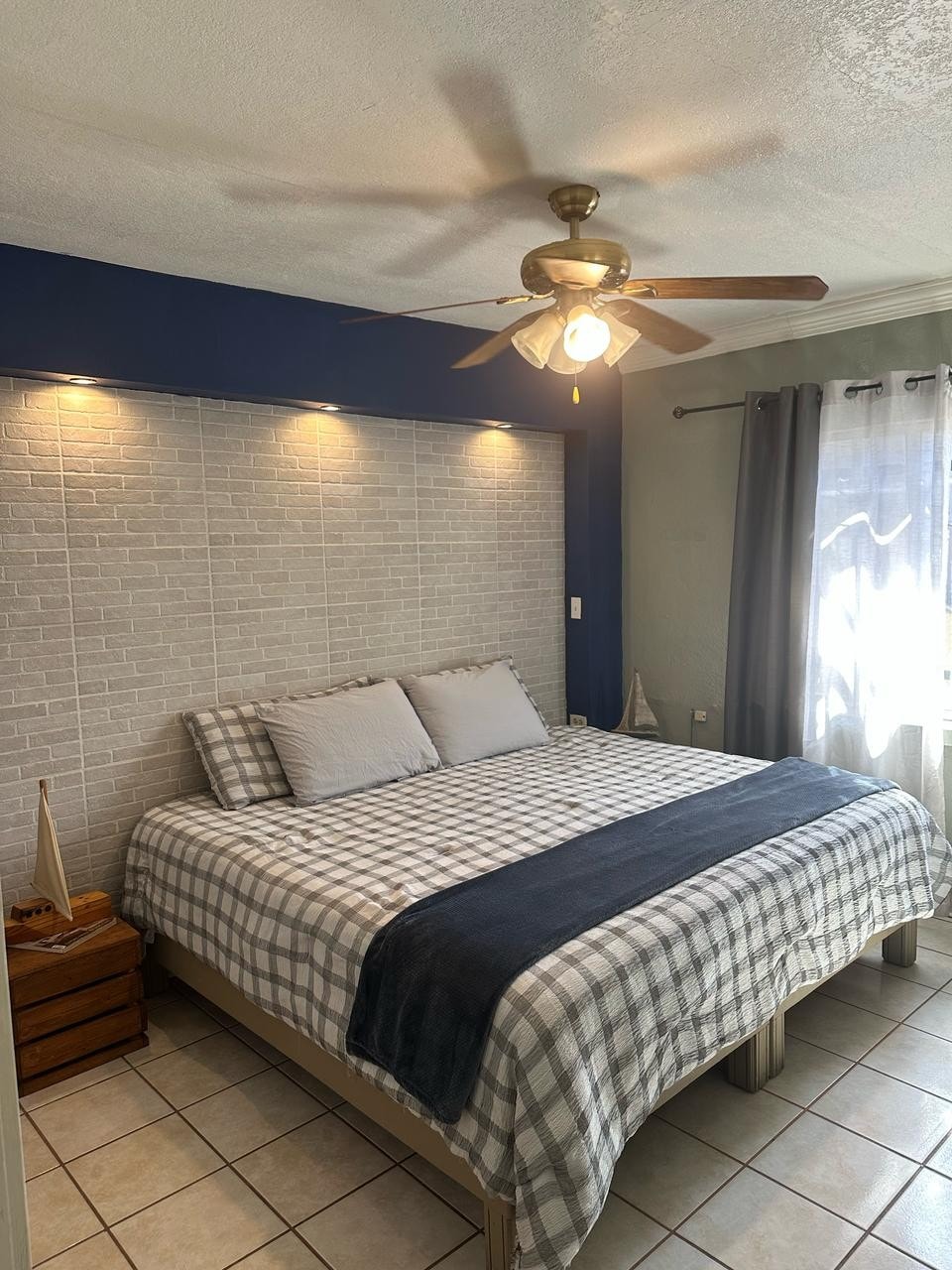 A spacious bedroom features a CalKing bed with a checkered blanket and gray pillows. Soft lighting is provided by a ceiling fan and wall sconces. Sheer curtains frame the window, allowing natural light to enhance the neutral tones of the room.