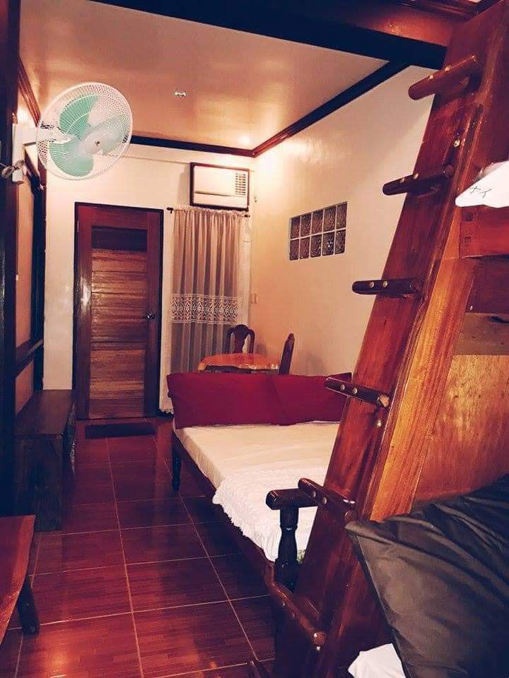 A cozy room features a spacious bed with crisp bedding positioned next to a wooden ladder leading to an upper sleeping area. A ceiling fan and wall-mounted air conditioning unit provide comfort, while a curtain drapes over a window that allows natural light to brighten the space.