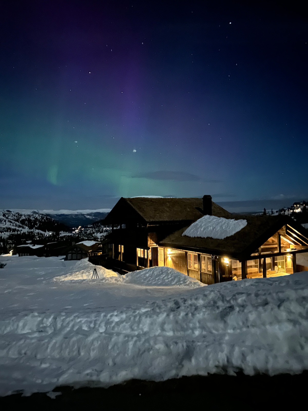 A mountain cabin is illuminated under a night sky, while the Northern Lights create a captivating display of green and purple hues overhead. Snow blankets the landscape surrounding the cabin, reflecting the soft glow from its exterior lighting.