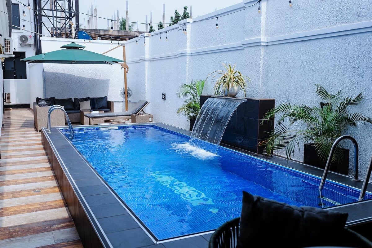 A refreshing swimming pool is highlighted by a waterfall feature, surrounded by tropical plants. A shaded seating area is positioned nearby, providing a comfortable spot for relaxation. The pool area is designed with sleek tiles and wooden decking, creating a serene atmosphere.