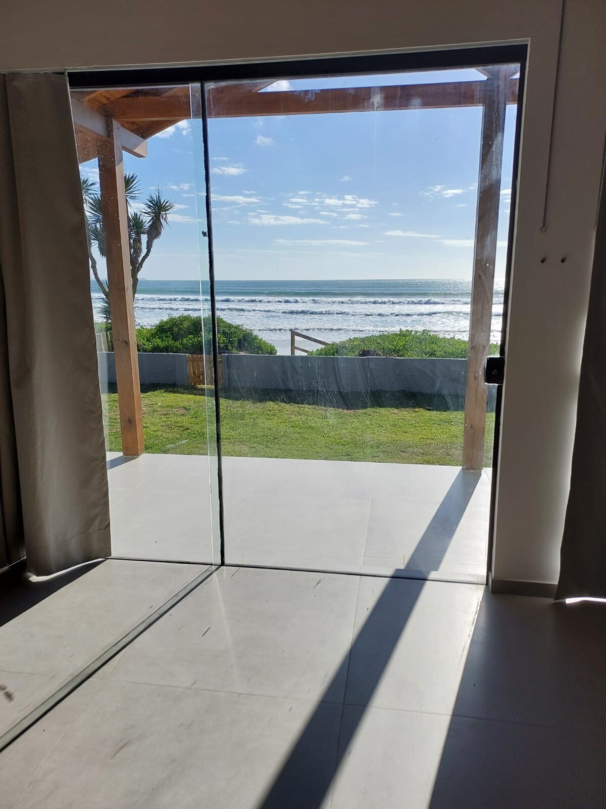 A glass door leads to an outdoor area, revealing a view of the ocean and grassy landscape. Sunlight filters through, creating soft shadows on the floor. The sky is clear with scattered clouds, enhancing the connection between indoor and outdoor spaces.