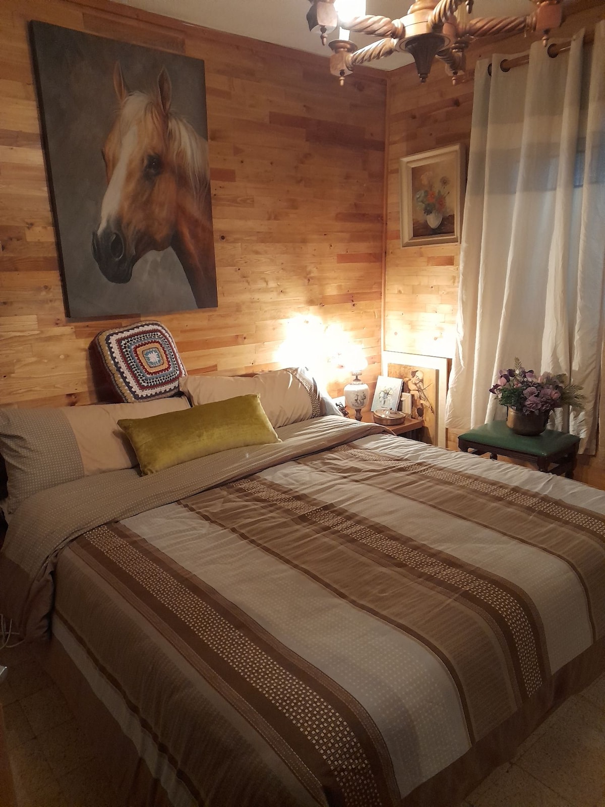 A cozy bedroom features warm wooden paneling and a large artwork of a horse above the bed. The bed is dressed with neutral-toned linens and a decorative pillow. A bedside lamp provides soft lighting, and a small table holds a vase of flowers.
