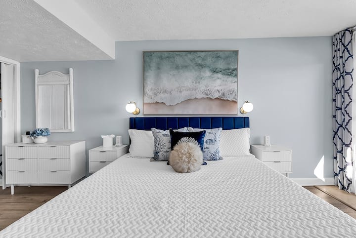 Relájate en un dormitorio principal tranquilo que cuenta con una lujosa cama tamaño king con ropa de cama blanca y fresca. El espacio se realza con una cabecera de terciopelo azul marino, mesitas de noche blancas modernas y una relajante paleta de colores costeros.