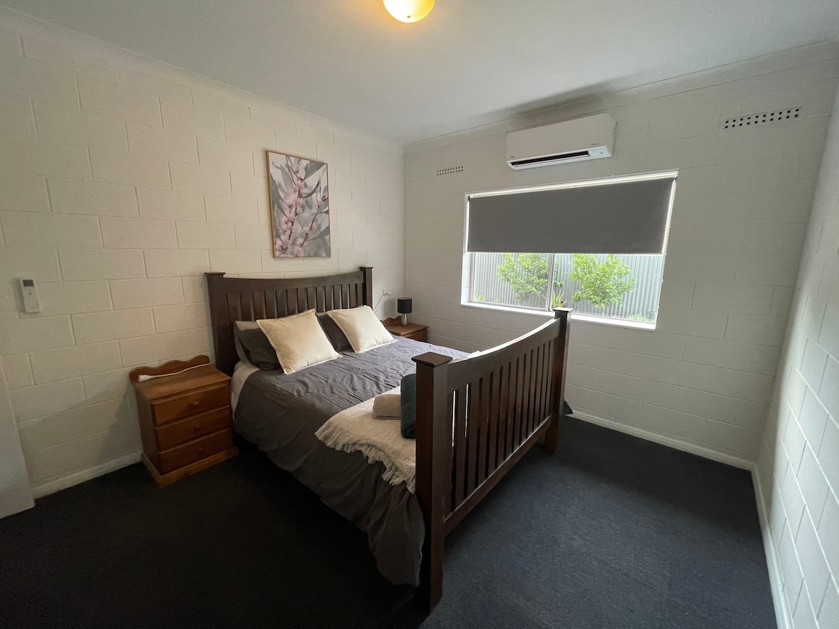 A comfortable bedroom features a wooden bed frame with bedding in neutral tones. A small bedside table sits next to the bed, and an air conditioning unit is mounted above the window, which is covered by a soft roller shade. Light enters through the window, illuminating the room.