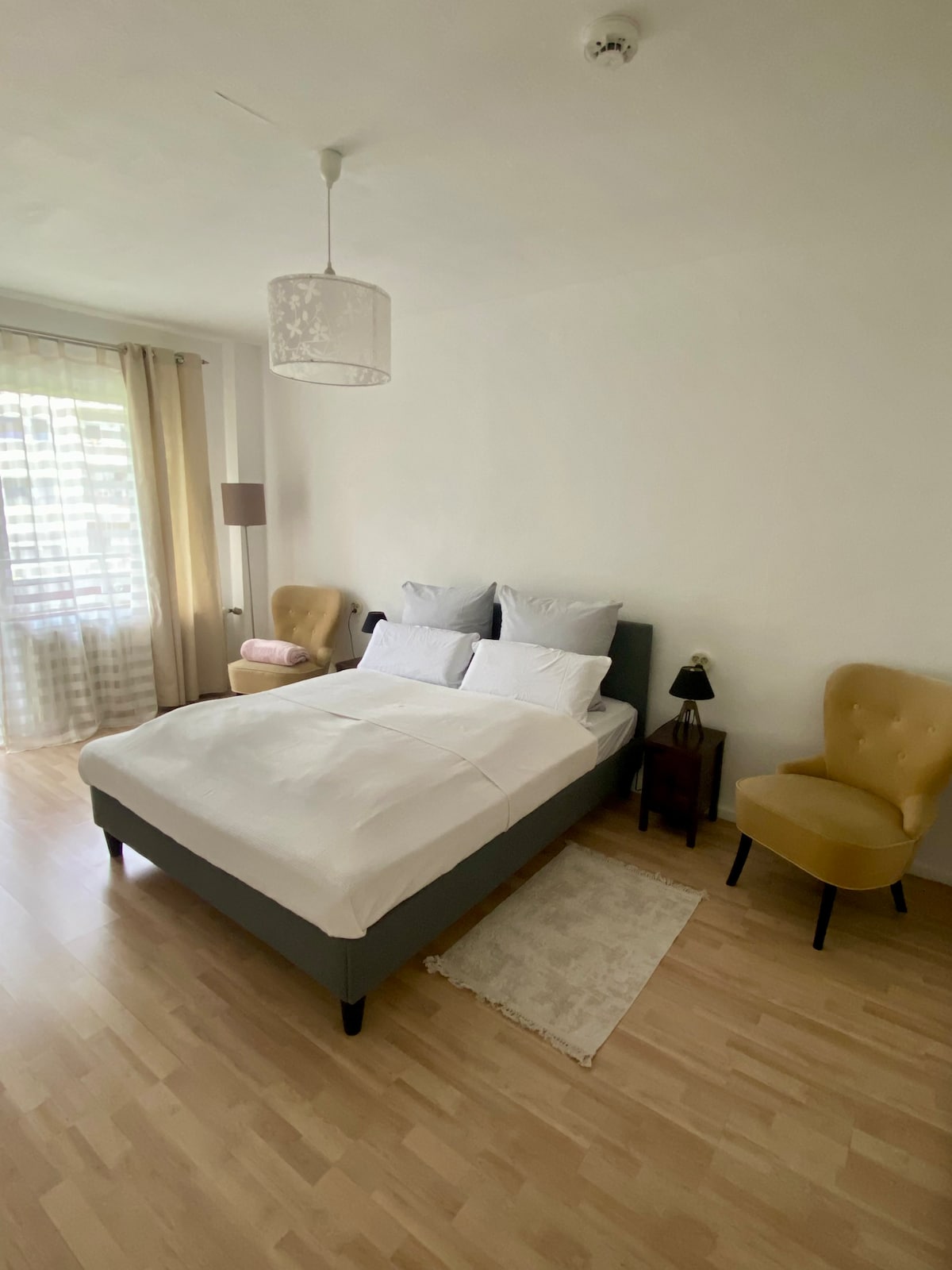 A bright bedroom features a spacious bed dressed in white linens, complemented by two yellow armchairs. Natural light filters in through sheer curtains, illuminating the wooden flooring. A small rug is placed beside the bed, and a bedside lamp adds a touch of warmth.