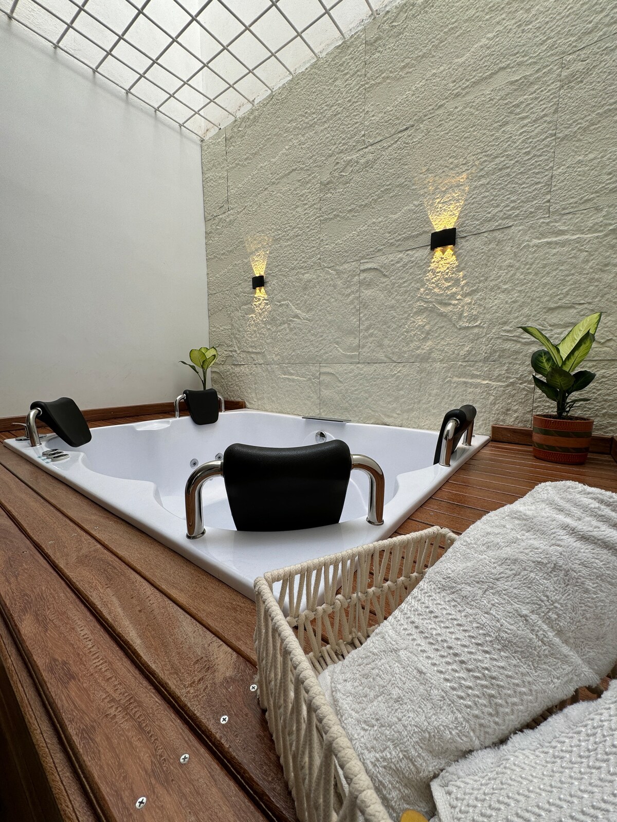 A modern jacuzzi is set within a serene space, surrounded by textured walls. Soft lighting emanates from wall sconces, and small potted plants add a touch of greenery. A wicker basket containing rolled towels rests nearby, enhancing the relaxation atmosphere.