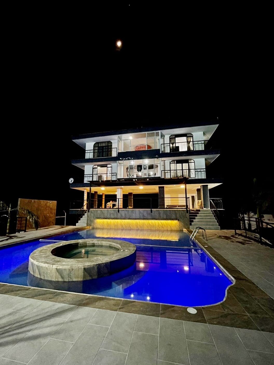 A modern three-story residence illuminated at night features a large swimming pool with a built-in hot tub. The sleek architecture includes spacious balconies offering views over the pool and surrounding area, with soft yellow lighting enhancing the inviting atmosphere.