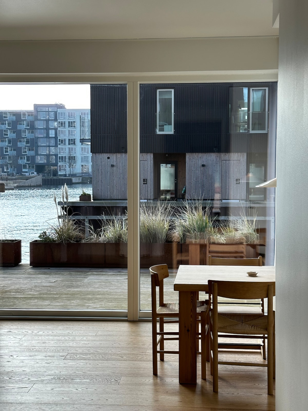 A dining area is positioned near large glass doors, offering views of the waterfront and adjacent modern buildings. A simple wooden table is surrounded by two chairs, with natural light enhancing the warmth of the wooden flooring. Potted plants adorn the exterior terrace.