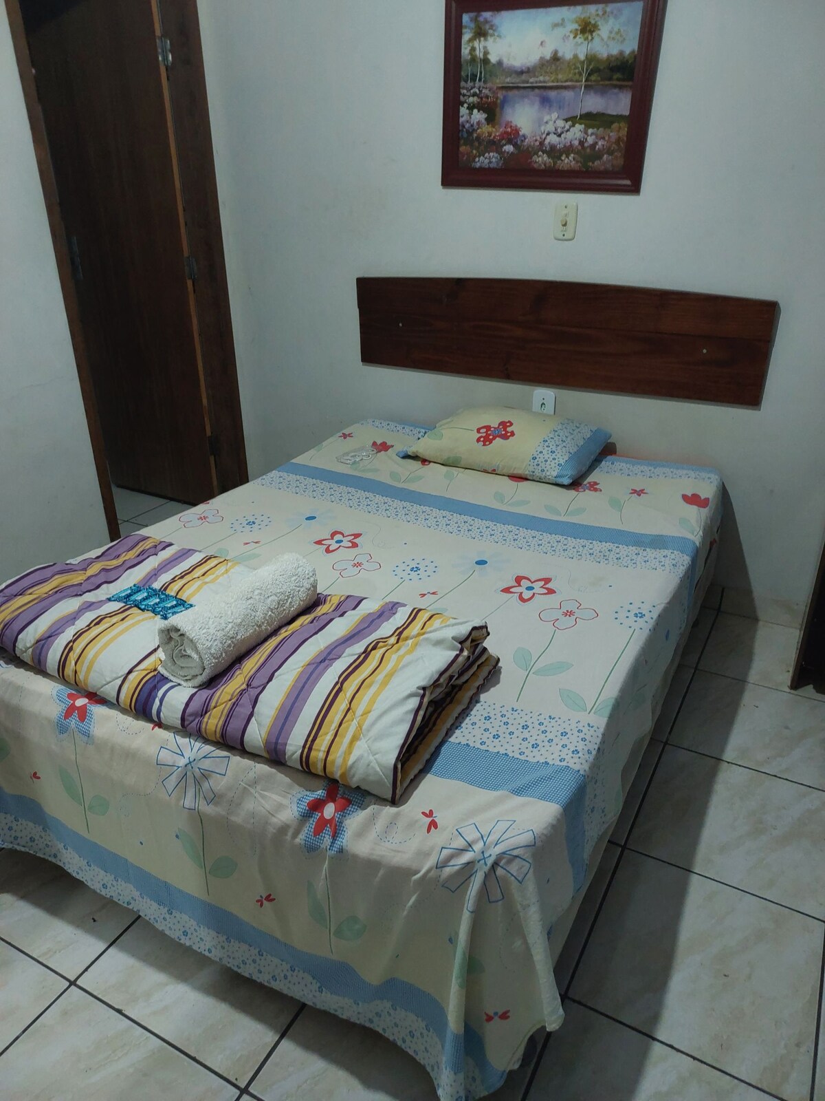 A modest sleeping area features a bed with a floral-patterned coverlet. Two pillows, one with a solid color and the other patterned, are placed at the head of the bed. A rolled towel is neatly arranged on the side, complemented by blankets folded at the foot.