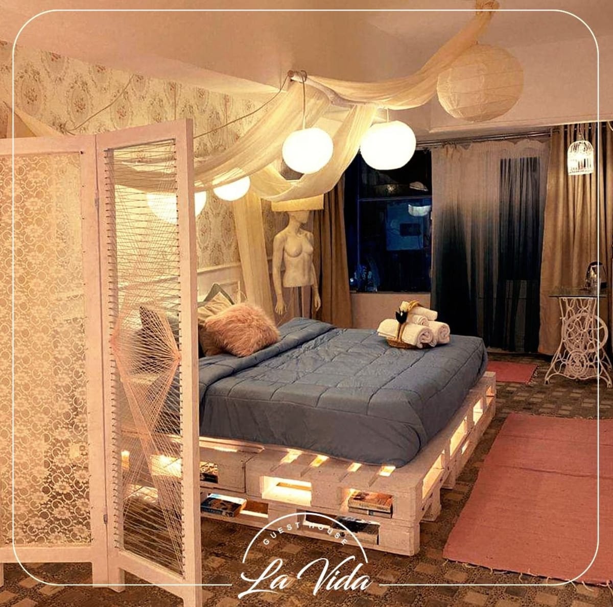 A creatively designed sleeping area features a spacious bed on a wooden platform, illuminated by soft hanging globes. Light wall art and a decorative partition contribute to the unique vibe, while light fabrics adorn the windows, allowing for gentle natural light to enter.