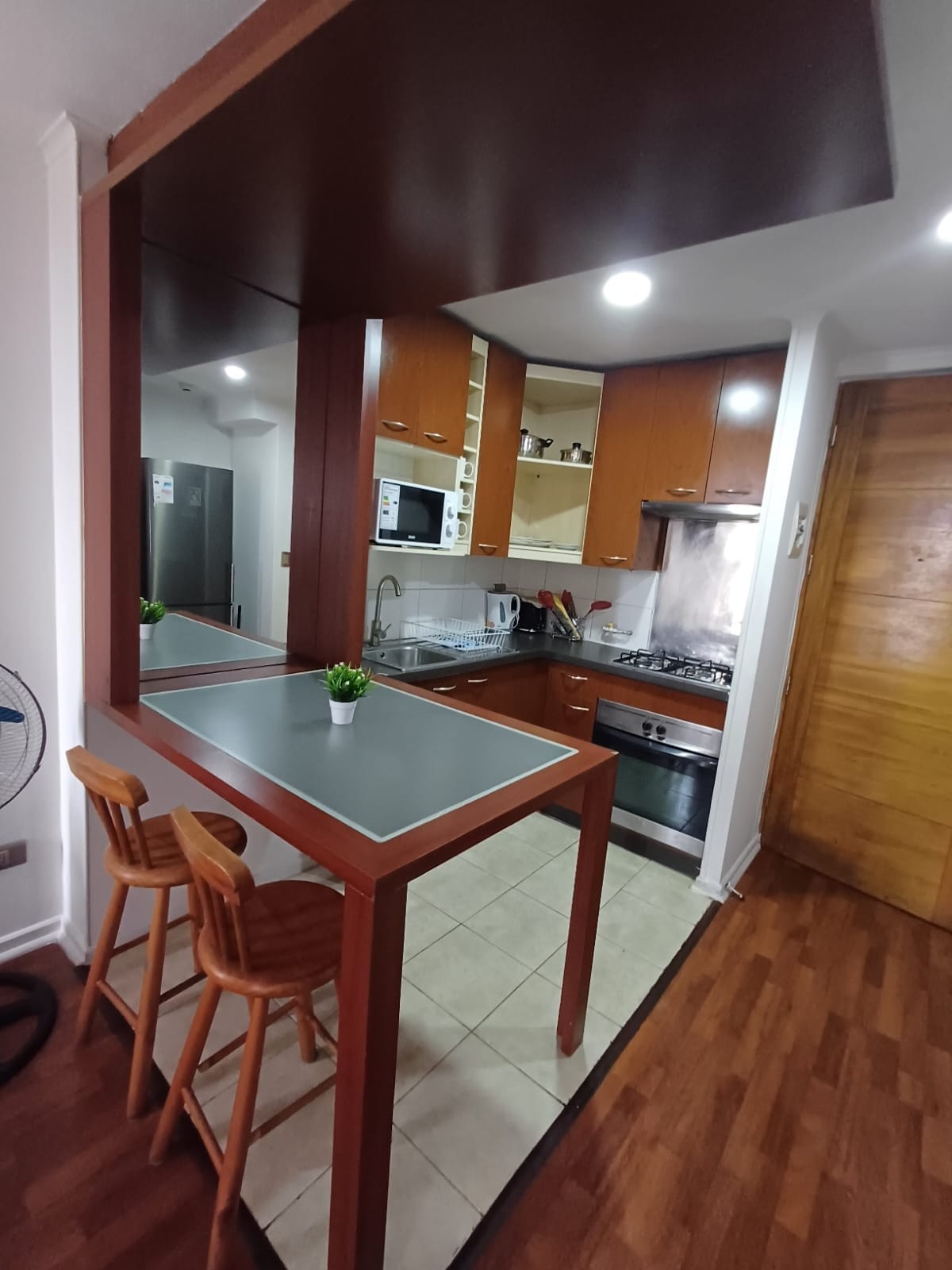 A well-organized kitchen is highlighted by warm wooden cabinetry and modern appliances. A compact breakfast bar with two wooden stools invites casual dining, while overhead shelving provides additional storage. Natural light illuminates the space through a small window, enhancing the functional layout.