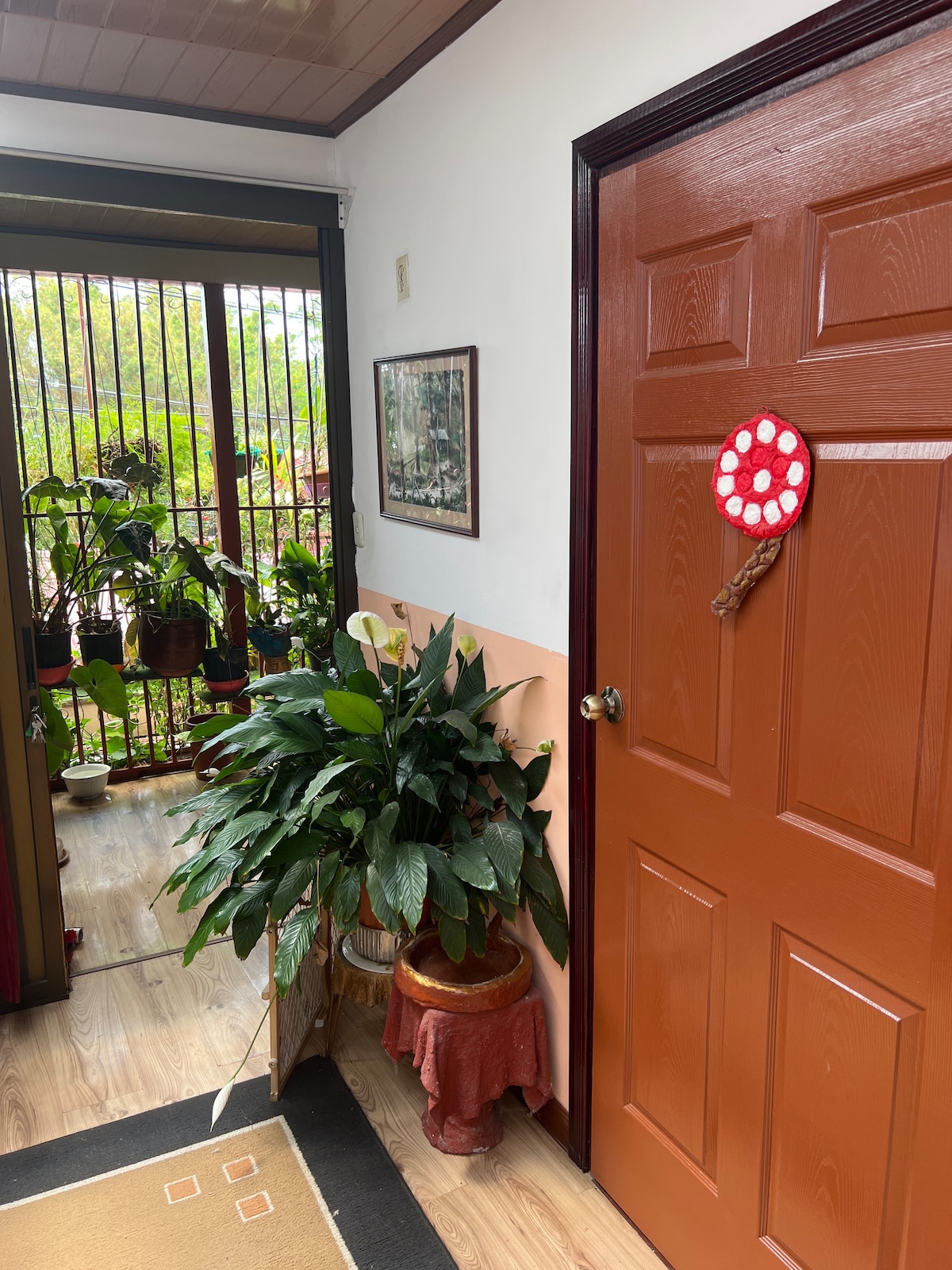 An entryway is depicted, featuring a warm-colored door adorned with a decorative piece. Lush green plants are arranged beside the door, complemented by a small table draped with fabric. Natural light filters through a window, creating a welcoming ambiance in the space.