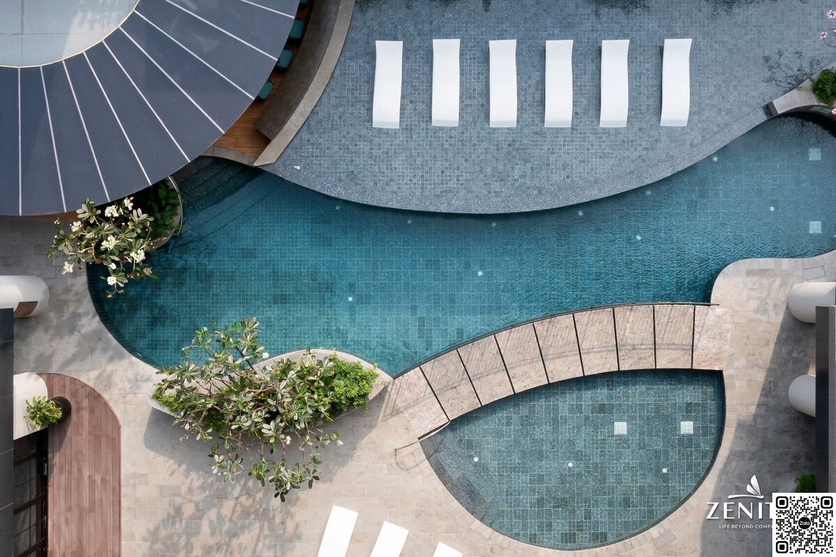 An aerial view showcases a uniquely designed swimming pool, featuring various shapes and levels. Surrounding the pool area are several lounge chairs neatly arranged on the stone pathway, while lush greenery adds natural elements to the scene.