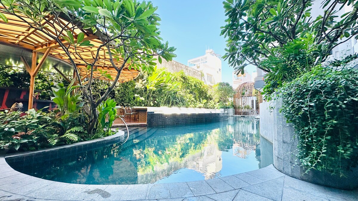 A serene swimming pool is surrounded by lush greenery and tropical plants, creating a peaceful oasis. The crystal-clear water reflects the blue sky, while wooden structures provide shaded seating areas. Decorative stone surrounds the pool, enhancing the tranquil ambiance.