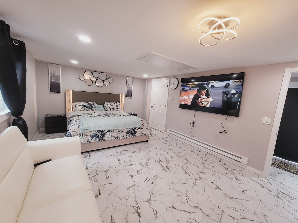 A spacious bedroom features a large television mounted on the wall opposite a comfortable sitting area. A floral-patterned bedspread decorates the bed, while elegant wall decor enhances the ambiance. Natural light is provided by the nearby window, covered with flowing black curtains.