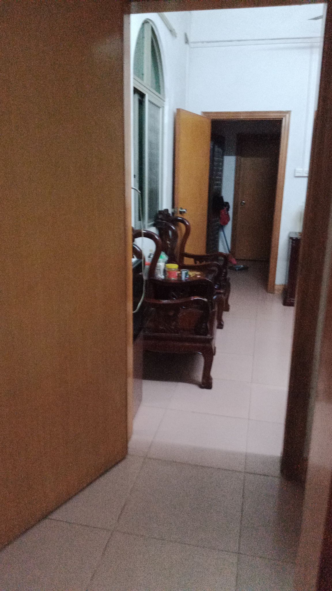 A narrow hallway is visible, leading to multiple wooden doors. Two ornate wooden chairs are positioned against the wall, with a small table in between. Natural light enters through a window, illuminating the space.