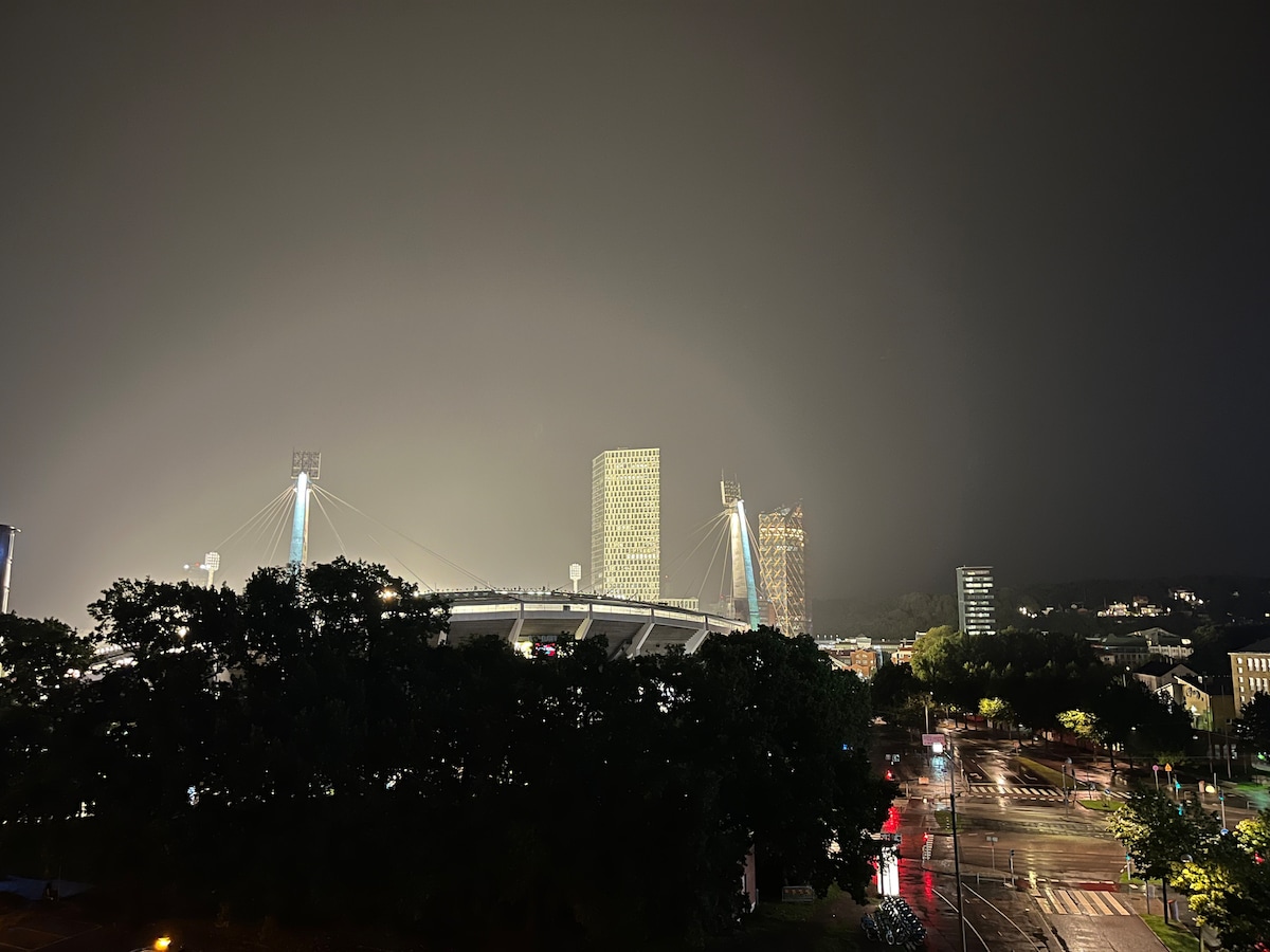 An evening cityscape is presented under a darkened sky, featuring the illuminated outlines of modern buildings. The nearby stadium is visible, along with treetops and a reflective wet pavement that enhances the urban atmosphere.