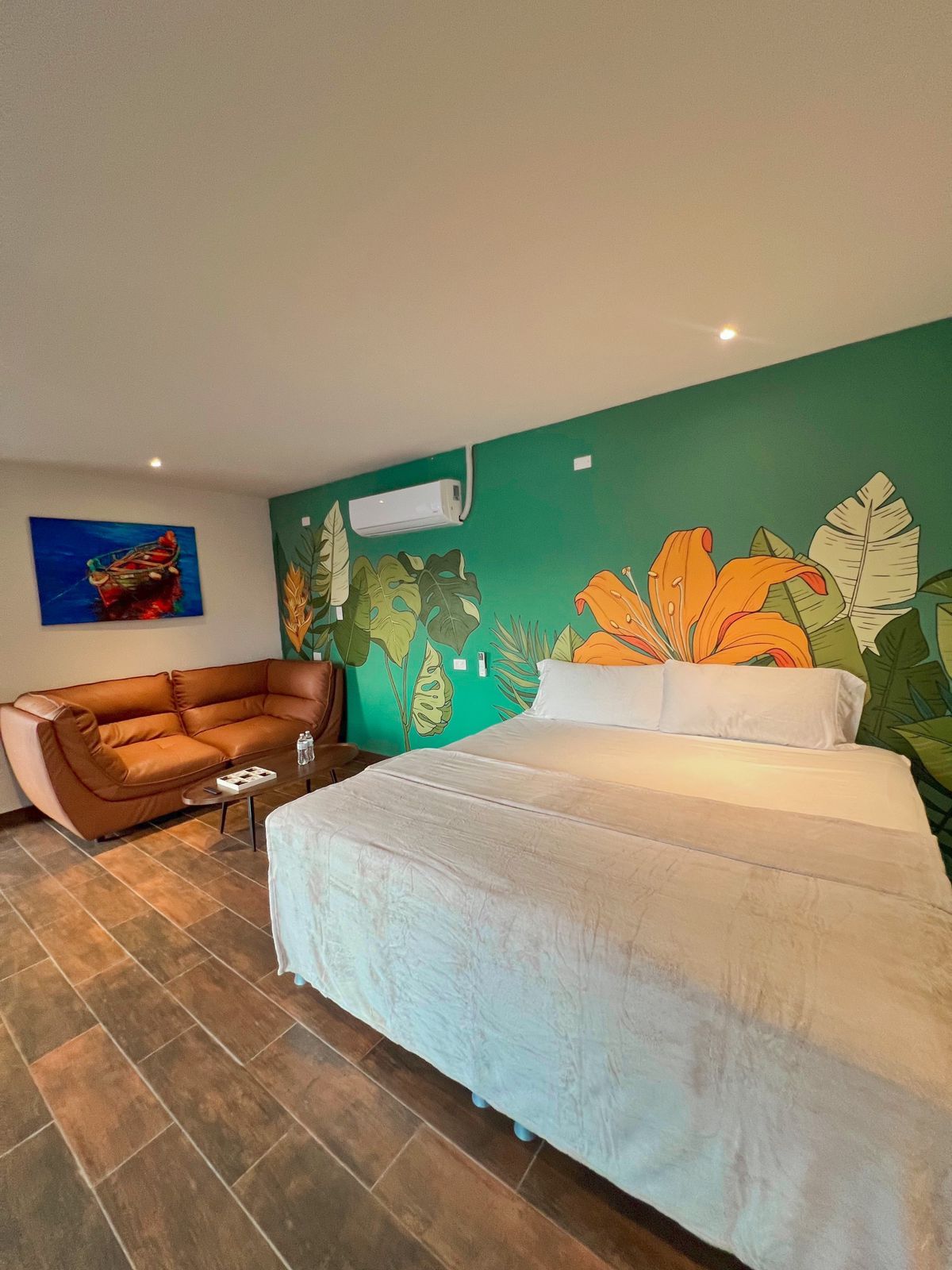 A colorful bedroom showcases a spacious bed with a light-colored coverlet, complemented by a cozy seating area with a brown couch. Vibrant murals of tropical plants adorn the walls, while soft lighting creates a relaxed ambiance.
