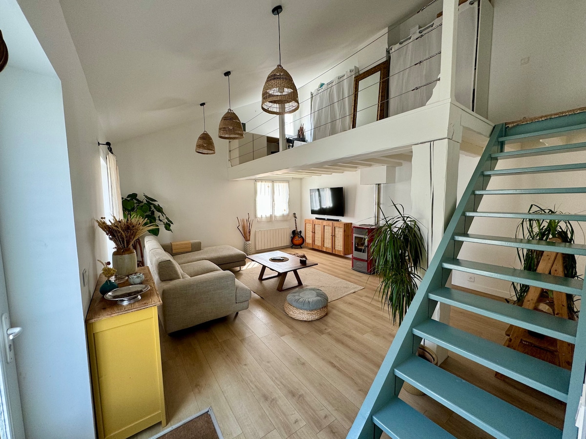 The living area features a spacious layout with a light-colored sofa and a central wooden coffee table. Decorative plants are placed throughout, while a staircase in a soft blue leads to the upper level. Natural light filters in through sheer curtains, enhancing the brightness.