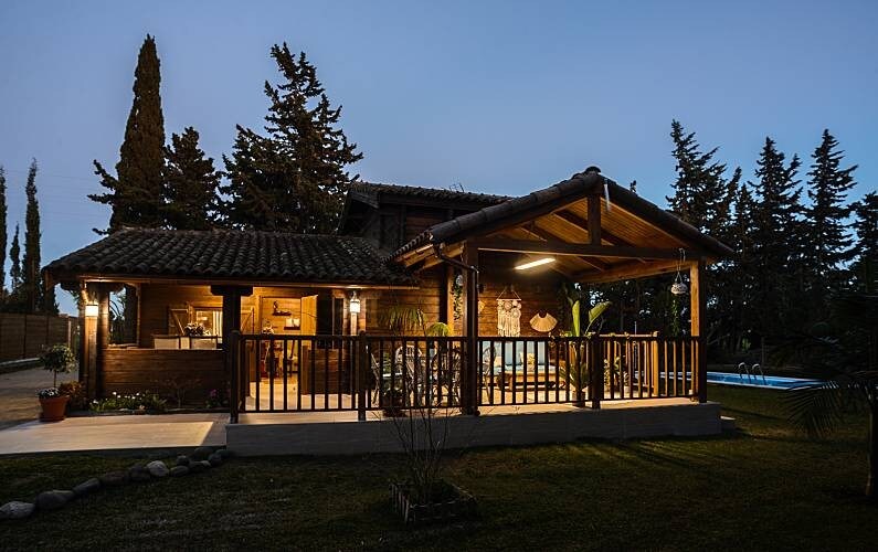 A rustic wooden house is showcased at dusk, with warm light illuminating the porch. Surrounding greenery and a pool area can be seen beyond the wooden railing, creating a serene outdoor atmosphere.