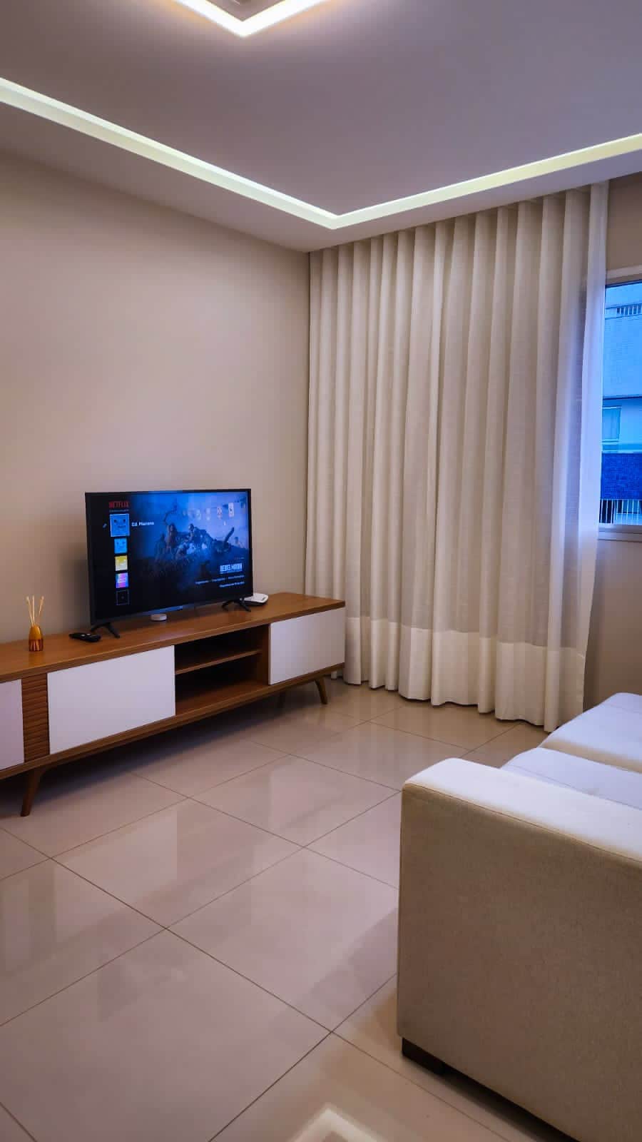 A cozy living area features a television mounted on a wooden stand, complemented by a light-colored sofa. Sheer curtains frame a window, allowing natural light to illuminate the room, while the tiled floor adds a sleek finish to the space.