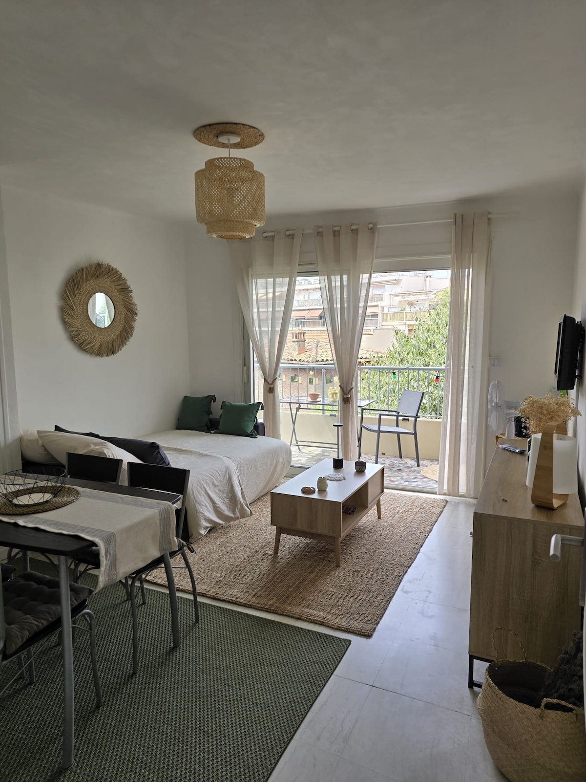 A bright living area features a spacious sofa, a low wooden coffee table, and a dining space with four chairs. Natural light fills the room through large glass doors leading to a private balcony. Light-colored curtains and decorative elements enhance the inviting atmosphere.