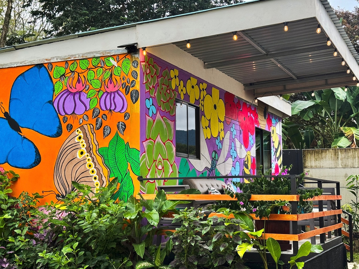A vibrant exterior of a tiny house is adorned with colorful murals depicting local flora and fauna. The covered terrace features wooden railings and is surrounded by lush greenery. A large window is framed by bright artwork, inviting a connection with nature.
