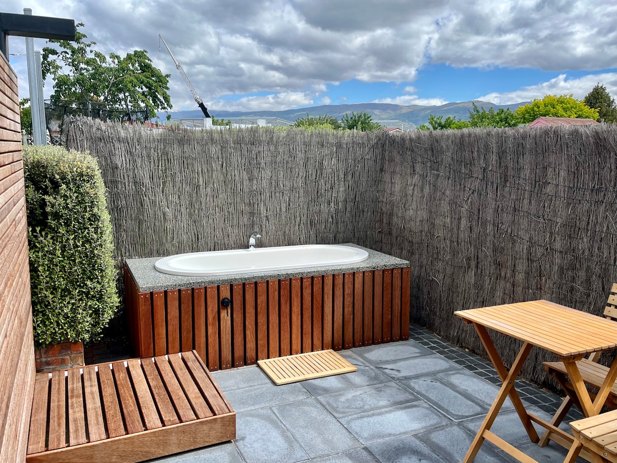 An outdoor patio area features a wooden hot tub surrounded by privacy fencing. A small wooden table and two chairs sit on the gray stone flooring, providing a cozy setting. Greenery and natural textures enhance the space, creating a peaceful retreat.
