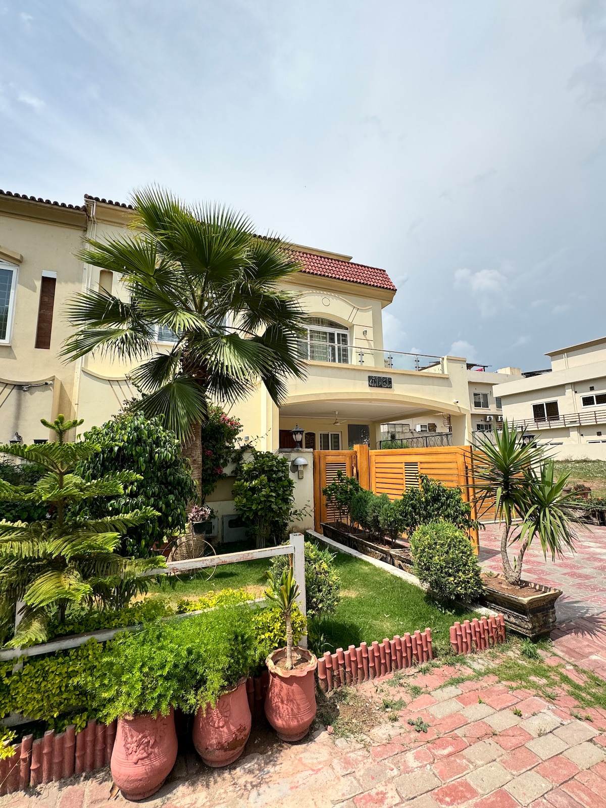 The exterior of the bungalow showcases a well-maintained garden featuring various green plants and a palm tree. A vibrant wooden gate opens to a welcoming entrance, with stone walkways leading through landscaped areas. The home features a cream-colored facade under a partly cloudy sky.