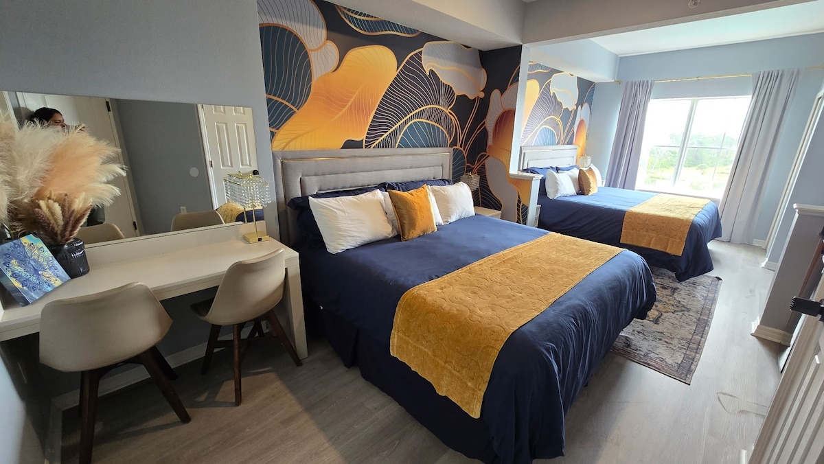 A spacious studio features two beds draped in navy blue linens with yellow accents. A decorative wall showcases a floral pattern. A desk and two chairs are positioned nearby, with natural light entering through large windows that are partially draped with light curtains.