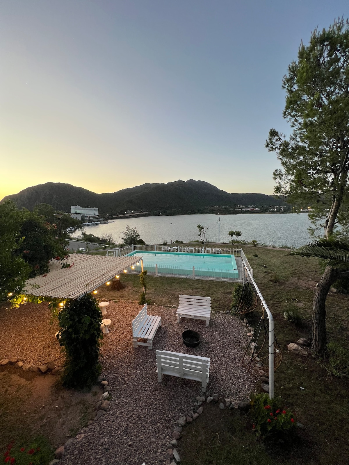 A serene outdoor area is seen with several white benches arranged on gravel. A private swimming pool glistens in the sunlight, while the surrounding landscape includes mountains and water. Soft lighting is provided by string lights above a shaded seating space.