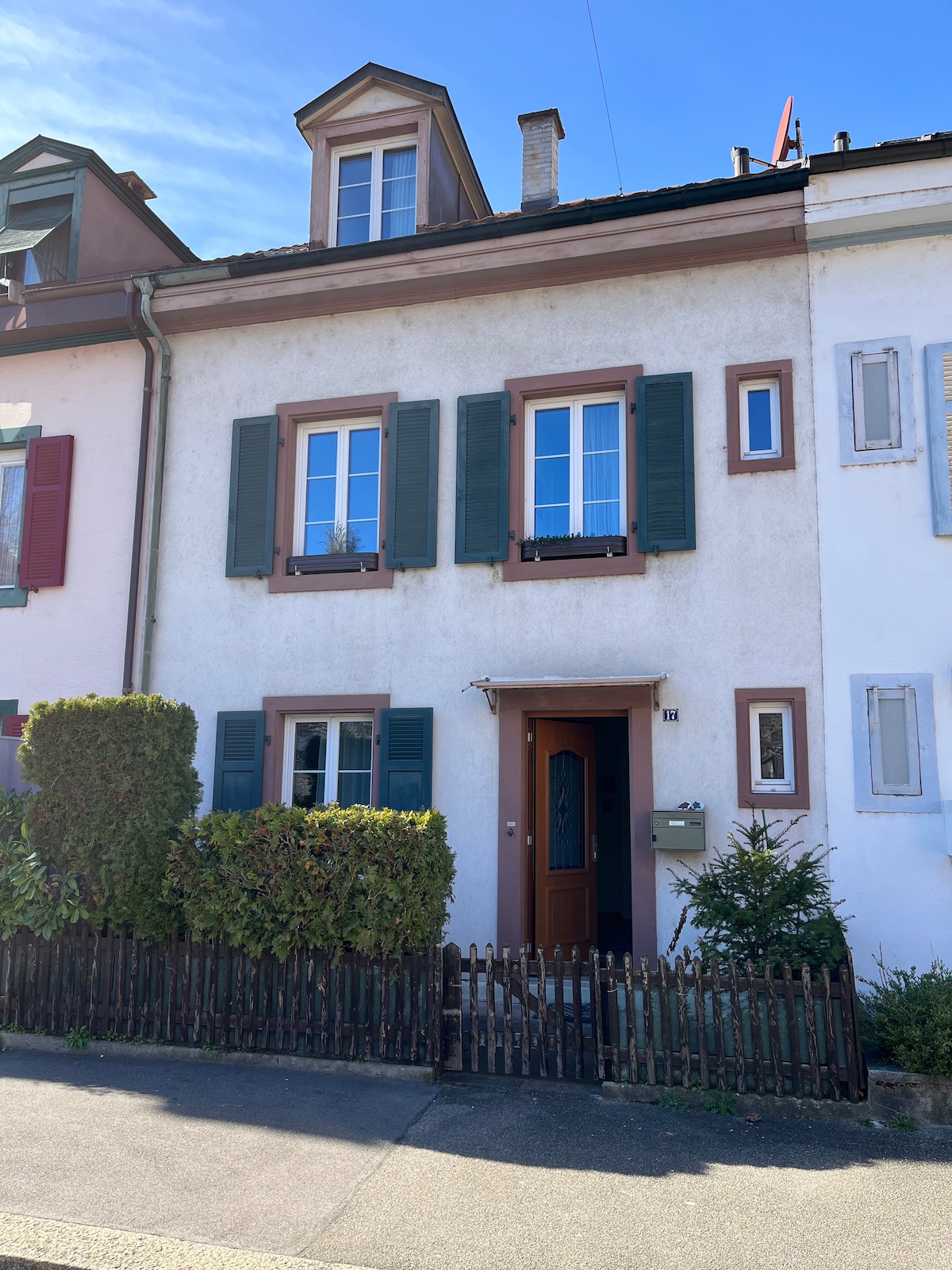 A charming two-story house is presented with a light-colored façade and shuttered windows. The front door is framed by a small porch, while the neat front yard features low hedges and a wooden picket fence. A sidewalk leads to the entrance, inviting guests inside.