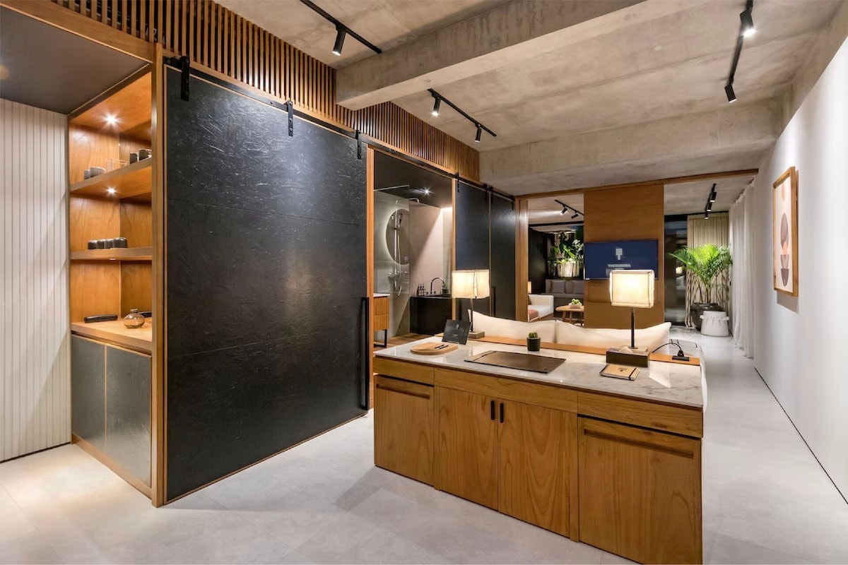 The studio features a contemporary open layout with wooden elements and a polished concrete ceiling. A well-lit area is furnished with a central countertop, decorative shelves, and various indoor plants, creating a modern yet welcoming environment.