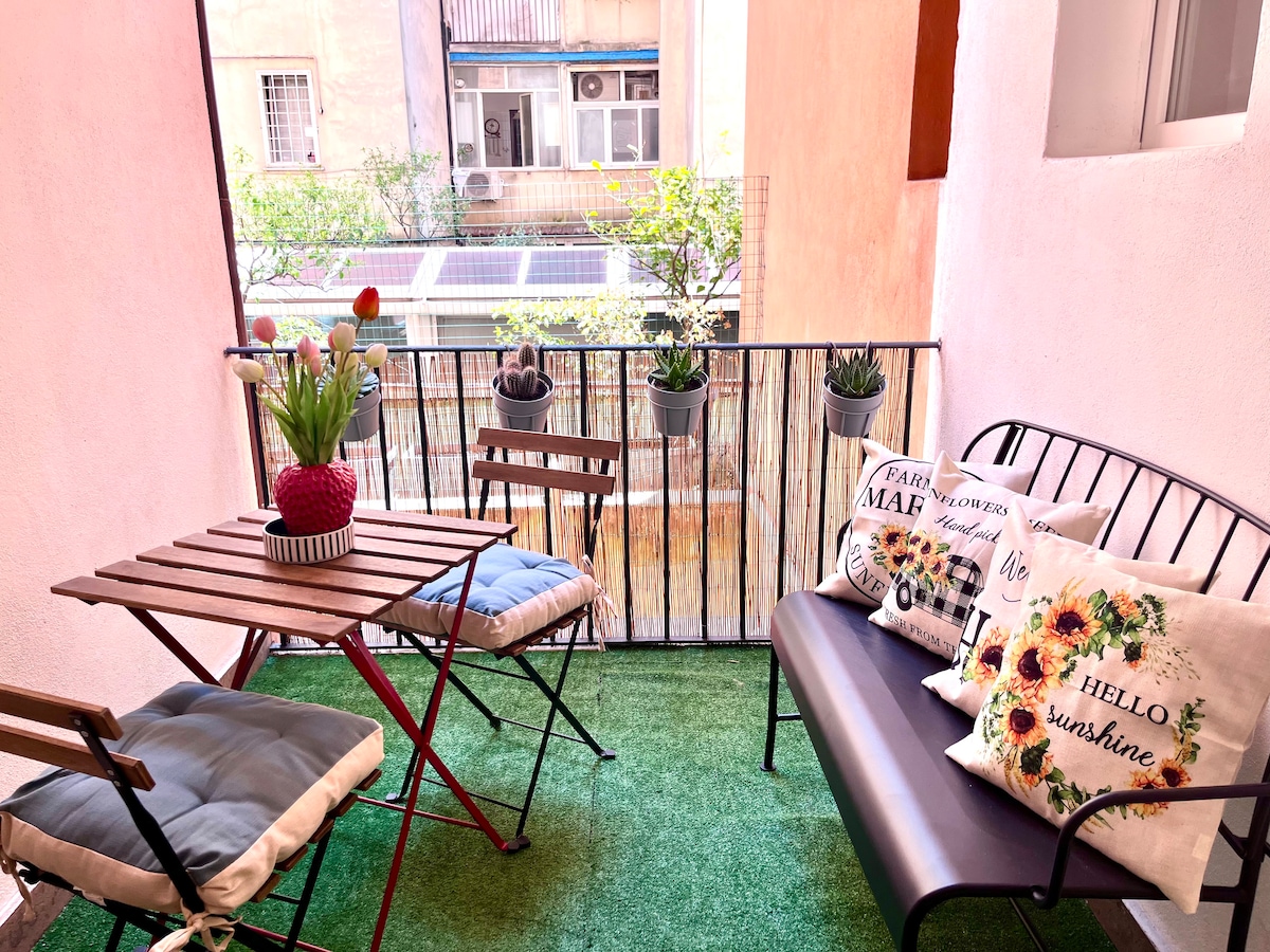 A cozy terrace is equipped with a small table and a bench, surrounded by potted plants and decorative cushions. The flooring is covered with green grass-like material, creating a relaxing area for guests to unwind outdoors.
