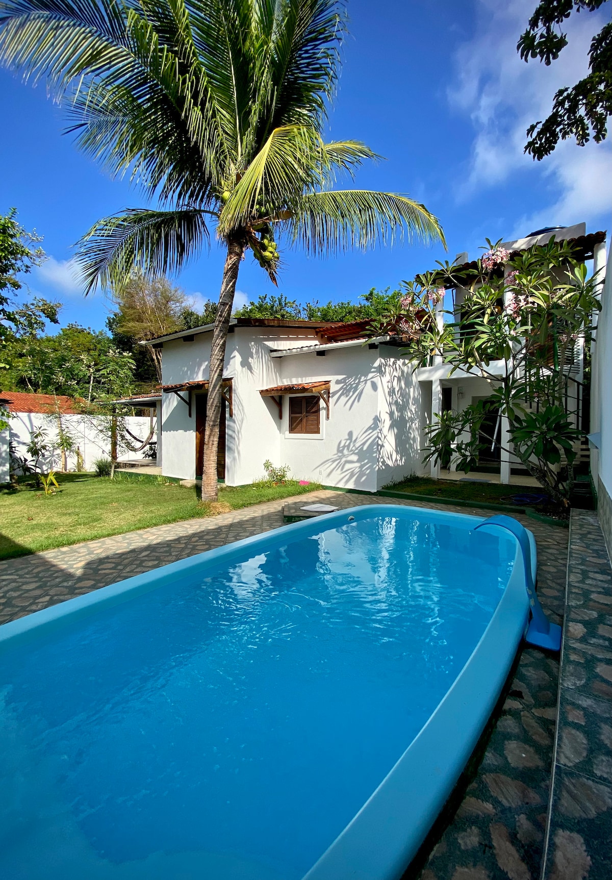 A private swimming pool is surrounded by lush greenery and palm trees, reflecting a bright blue sky. The pool is complemented by a patterned stone pathway leading to a charming single-story home with wooden windows, providing a serene outdoor space for relaxation.