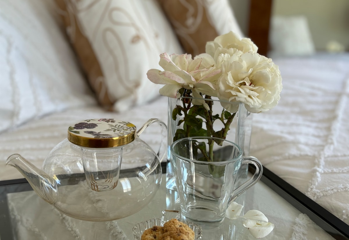 A clear glass teapot is placed on a reflective tray alongside a delicate glass cup. Fresh white flowers are arranged in a vase, adding a touch of nature. Light-colored linens cover the spacious bed in the background, complementing the soft decor.