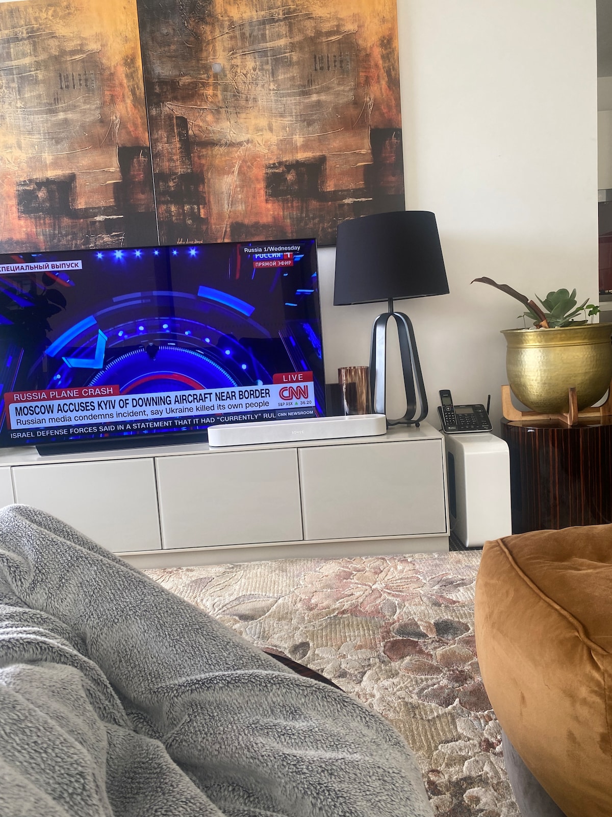 A stylish living area is showcased, featuring a large television displaying a news broadcast. A modern lamp stands beside the TV on a sleek white unit. A plush brown chair and woven rug add warmth to the space, complemented by a decorative potted plant.